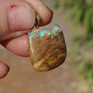 Puede incluir: Un colgante de ópalo marrón y dorado con una anilla de plata. El ópalo tiene un brillo iridiscente verde y azul.