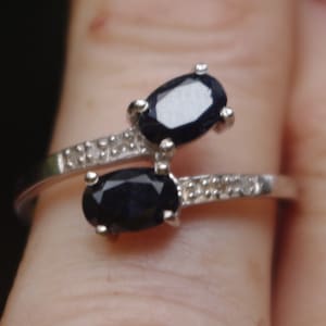 Puede incluir: Un anillo de plata con dos piedras preciosas negras de forma ovalada engastadas en la banda. El anillo está decorado con pequeñas piedras transparentes.