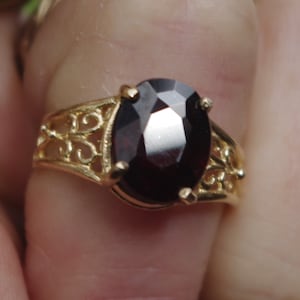Puede incluir: Un anillo de oro con una piedra ovalada de color rojo oscuro engastada en un diseño de filigrana.