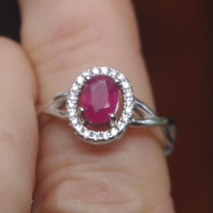 May include: A silver ring featuring a pink oval ruby gemstone set within a halo of smaller white gemstones.