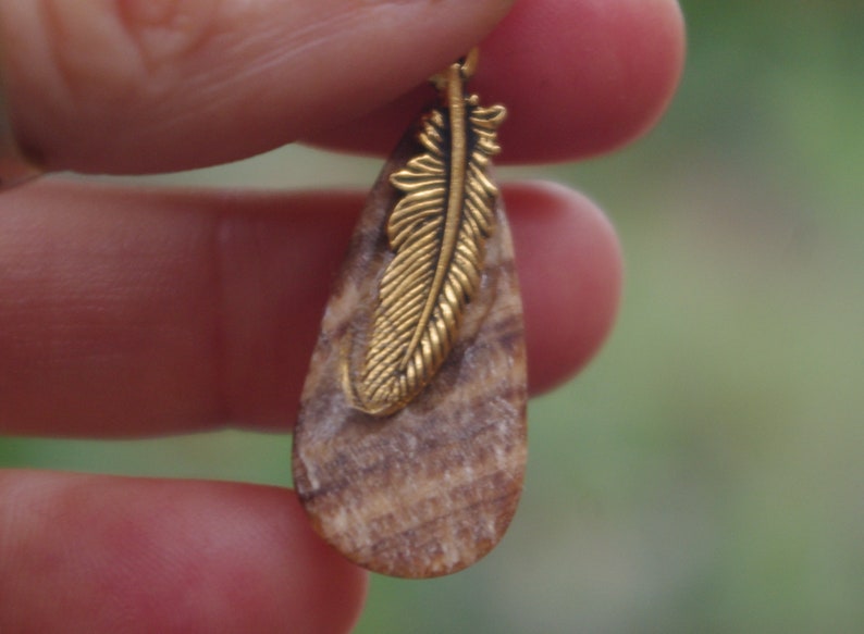 May include: A teardrop-shaped pendant made of wood with a gold-toned feather design.