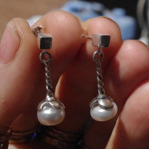 Puede incluir: Un par de pendientes de plata con un pequeño dije cuadrado y una perla colgante. Los pendientes tienen una cadena de plata trenzada que conecta el dije con la perla.