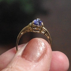 Puede incluir: Un anillo de oro con una sola piedra preciosa morada facetada en forma de pera, montada en un engaste de garras. La banda del anillo presenta un dise&ntilde;o decorativo calado con recortes en los laterales que conducen al engaste central.
