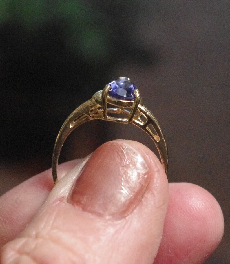Puede incluir: Un anillo de oro con una sola piedra preciosa morada facetada en forma de pera, montada en un engaste de garras. La banda del anillo presenta un dise&ntilde;o decorativo calado con recortes en los laterales que conducen al engaste central.