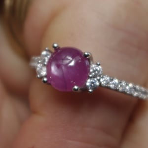 May include: A silver ring featuring a pink oval gemstone surrounded by smaller clear gemstones. The ring has a delicate band with a row of clear gemstones.