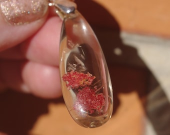 May include: A clear glass pendant with a silver bail, containing small red and orange crystals.