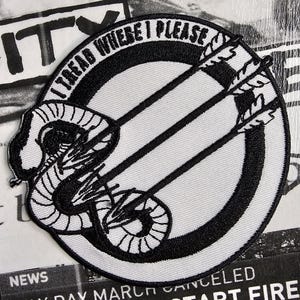 May include: A circular patch with the text "I TREAD WHERE I PLEASE" in a black border. The patch features a black snake and arrows design on a white background. The patch is on a background of black and white printed material.