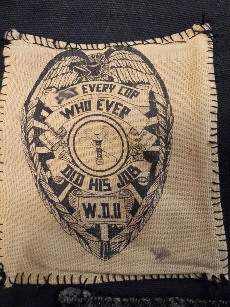 May include: A tan canvas patch with a black and white image of a police badge. The badge has an eagle, a scroll with the text "Every Cop Who Ever Did His Job", and the acronym "W.D.J."