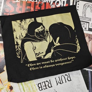 May include: Black square textile with a gold illustration of two figures, one masked. Text reads "Then we must do without hope. There is always vengeance!" surrounded by concert posters.