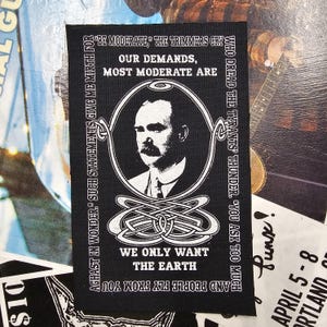 May include: Black and white patch featuring a portrait of a man with a moustache, encircled by text. The patch includes the phrase "WE ONLY WANT THE EARTH" and other quotes. The design incorporates Celtic knotwork and a vintage style.