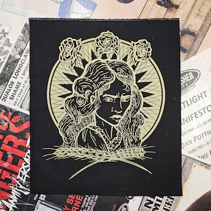 May include: Black patch with a gold illustration of a woman with roses and wheat. The design is set within a circular frame. The patch has a raw edge. The image is reminiscent of a vintage design.