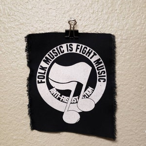 May include: A black fabric patch with white text and a musical note design. The text reads "FOLK MUSIC IS FIGHT MUSIC" and "ANTI-FASCIST ACTION". The patch is held by a black binder clip.