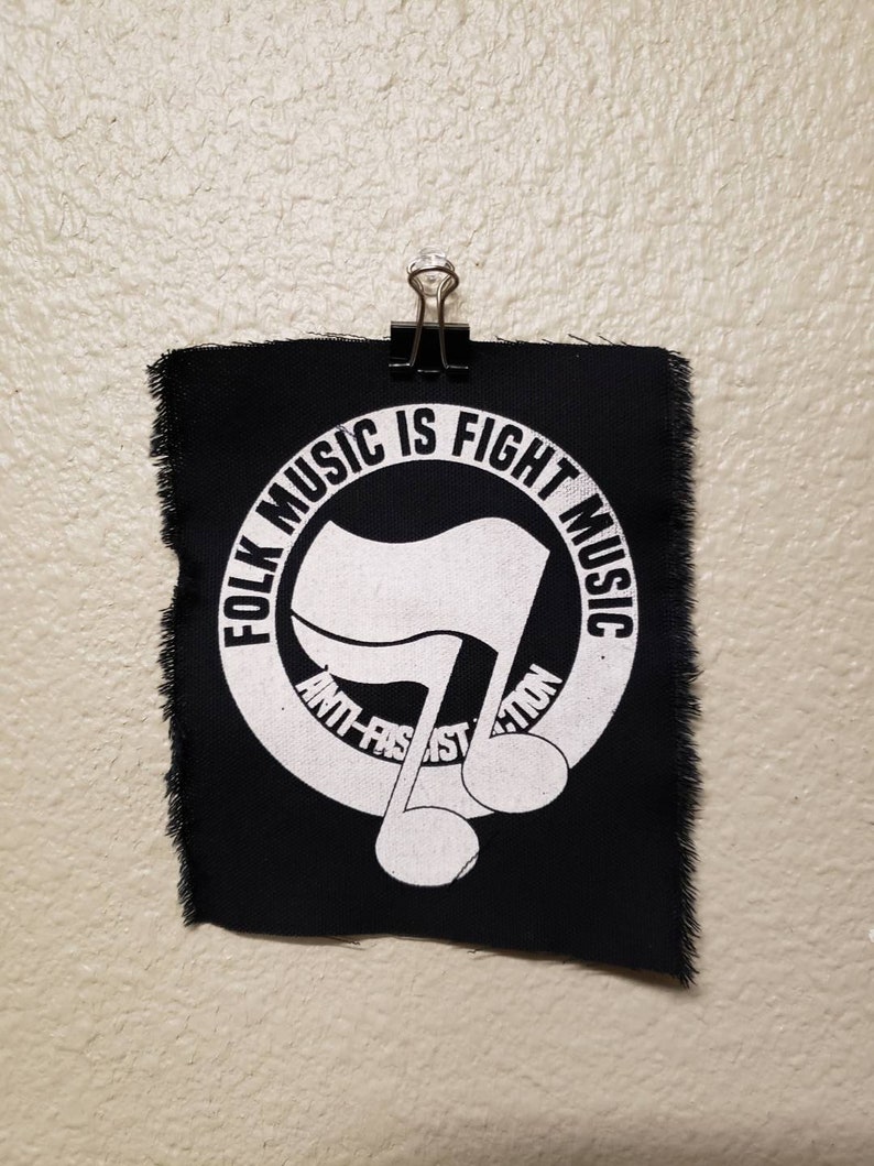 May include: A black fabric patch with white text and a musical note design. The text reads "FOLK MUSIC IS FIGHT MUSIC" and "ANTI-FASCIST ACTION". The patch is held by a black binder clip.
