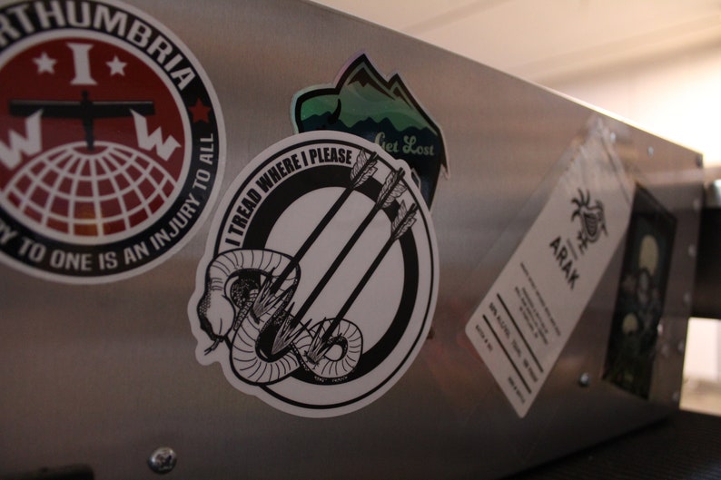 May include: A collection of stickers on a brushed metal surface. Stickers include a red and white circular design with text, a mountain scene, and a black and white design with arrows and a snake. One sticker reads "I TREAD WHERE I PLEASE."
