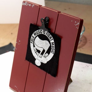 May include: A red wooden clipboard with a black metal clip holding a black patch. The patch features the text "FOLK MUSIC IS FIGHT MUSIC" in a circle around a stylized flag and musical notes.