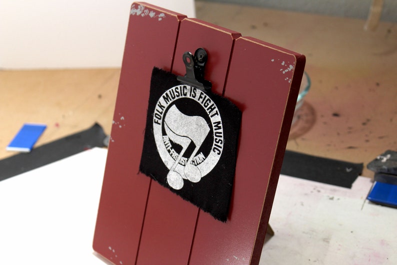May include: A red wooden clipboard with a black metal clip holding a black patch. The patch features the text "FOLK MUSIC IS FIGHT MUSIC" in a circle around a stylized flag and musical notes.