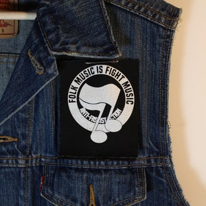 May include: A blue denim vest with a black patch featuring the text "FOLK MUSIC IS FIGHT MUSIC" in a circular design. The patch also includes a stylized musical note and the words "ANTI-FASCIST ACTION".