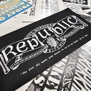 Pode incluir: Impressão retangular preta com a palavra "Republic" em branco, arqueada sobre uma faixa. A impressão inclui texto adicional e um brasão, com a citação "Os grandes só parecem grandes porque estamos de joelhos; levantemo-nos."