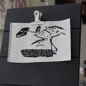May include: A black and white printed fabric patch featuring a skeletal creature and the text "I did not think myself frail, until facing the winds of your winter."