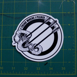 May include: A circular sticker with a black and white design. The design features a coiled snake, three arrows, and the text "I TREAD WHERE I PLEASE" in a circular pattern. The sticker is on a green surface.
