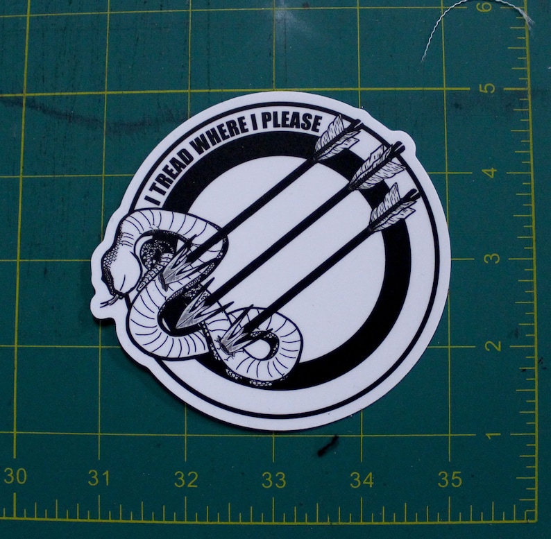 May include: A circular sticker with a black and white design. The design features a coiled snake, three arrows, and the text "I TREAD WHERE I PLEASE" in a circular pattern. The sticker is on a green surface.