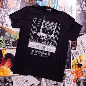 May include: Black t-shirt with a white graphic design featuring a building and text. The text reads "THIS IS NOT A PLACE OF HONOR" and "NOTHING VALUED IS HERE". Below the text are six stylized faces.