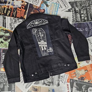 May include: Black denim jacket with two patches on the back. The top patch has a skull design, and the bottom patch says "Nelson's Blood" with a coffin illustration. The jacket is buttoned and has two chest pockets.