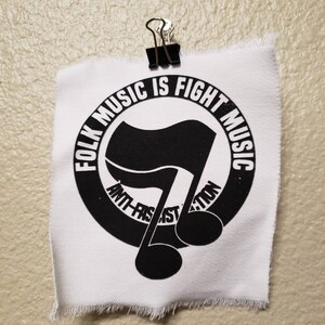 May include: A white fabric square with a black graphic and text, held by a black binder clip. The graphic is a musical note with a flag design inside a circle. Text reads "FOLK MUSIC IS FIGHT MUSIC" and "ANTI-FASCIST ACTION."