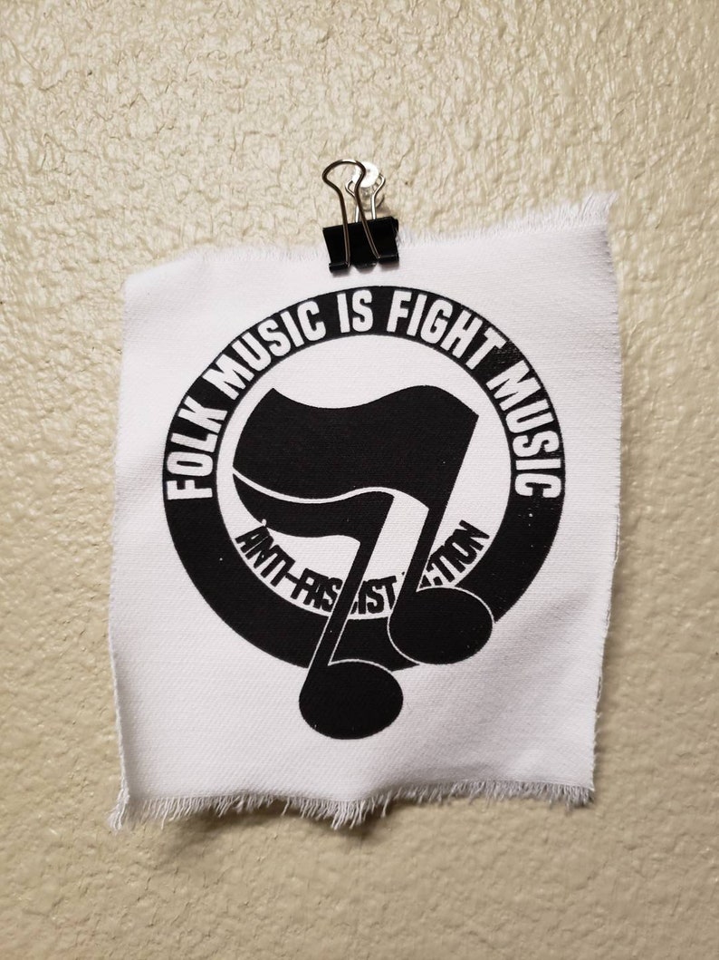 May include: A white fabric square with a black graphic and text, held by a black binder clip. The graphic is a musical note with a flag design inside a circle. Text reads "FOLK MUSIC IS FIGHT MUSIC" and "ANTI-FASCIST ACTION."