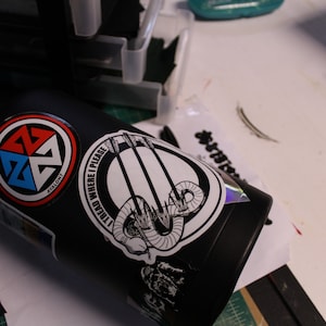 May include: Black water bottle with multiple stickers. One sticker has a red, blue, and white design with the word "Avalon". Another sticker has the text "I TREAD WHERE I PLEASE" with a graphic of arrows and a snake.