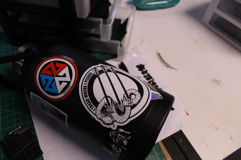 May include: Black water bottle with multiple stickers. One sticker has a red, blue, and white design with the word "Avalon". Another sticker has the text "I TREAD WHERE I PLEASE" with a graphic of arrows and a snake.