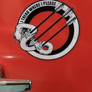 May include: A circular sticker with a black and white graphic of a coiled snake pierced by three arrows. The sticker has the text "I TREAD WHERE I PLEASE" in a circular pattern around the design. The sticker is on a red surface.