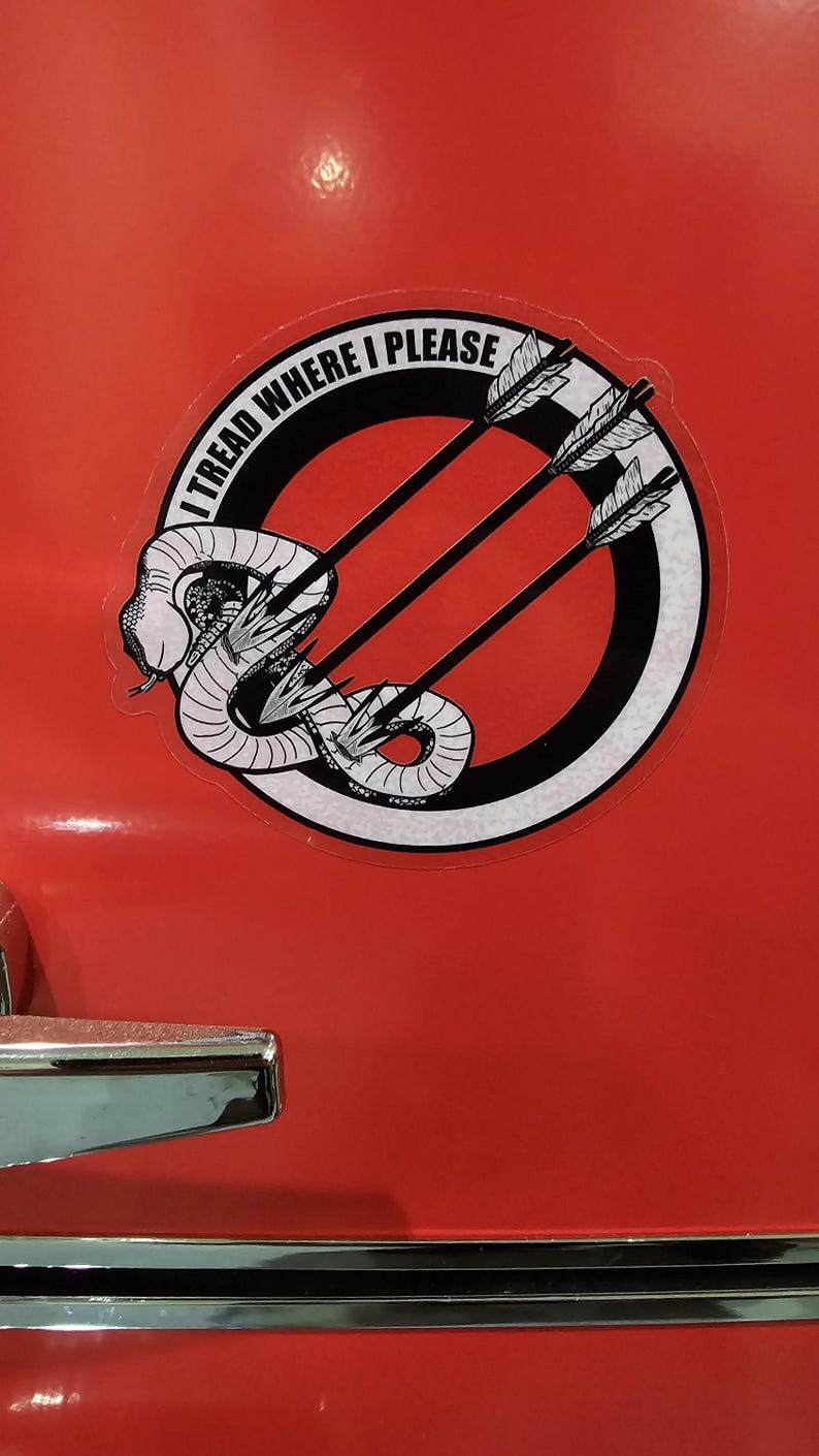 May include: A circular sticker with a black and white graphic of a coiled snake pierced by three arrows. The sticker has the text "I TREAD WHERE I PLEASE" in a circular pattern around the design. The sticker is on a red surface.