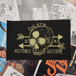May include: Black rectangular patch with gold text and a skull graphic. The text reads "DEATH IS AN ACCEPTABLE OUTCOME" with arrows pointing to the skull. The patch is surrounded by various posters and flyers.