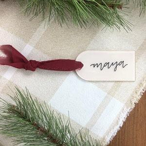 May include: A cream-colored place card with the name "maya" in black script, tied with a burgundy ribbon. The card is set on a beige and white plaid tablecloth, with pine branches in the background.