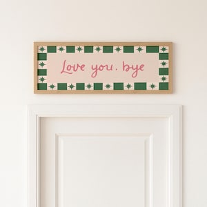 Puede incluir: Una obra de arte mural rectangular enmarcada con el texto "Love you. bye" en escritura rosa. La obra de arte tiene un fondo rosa claro con un borde verde y blanco de estrellas y cuadrados, y un marco de madera natural.