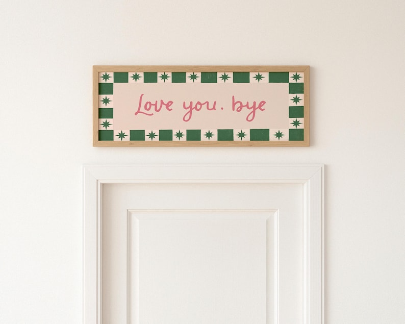Puede incluir: Una obra de arte mural rectangular enmarcada con el texto "Love you. bye" en escritura rosa. La obra de arte tiene un fondo rosa claro con un borde verde y blanco de estrellas y cuadrados, y un marco de madera natural.
