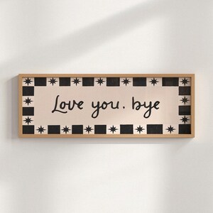 Puede incluir: Cartel rectangular de madera con las palabras "Love you, bye" en escritura negra. El cartel tiene un fondo color crema con un borde de cuadrados negros y estrellas alternas. El marco es de color marr&oacute;n claro.