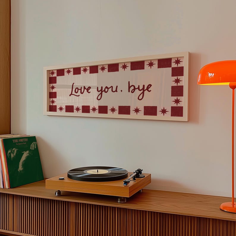 Puede incluir: Una obra de arte enmarcada con el texto "Love you. bye" en un estilo manuscrito, rodeada por un borde rojo y blanco. Un tocadiscos de madera y una pila de discos de vinilo est&aacute;n en un mueble de madera debajo.