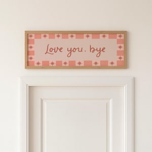 Puede incluir: Un letrero rectangular con marco de madera con las palabras "Love you. bye" en un estilo manuscrito. El letrero tiene un fondo rosa con un borde de cuadrados rosas y formas de estrella. El letrero est&aacute; montado sobre una puerta blanca.