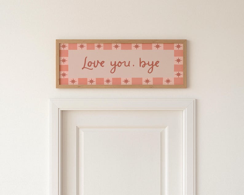 Puede incluir: Un letrero rectangular con marco de madera con las palabras "Love you. bye" en un estilo manuscrito. El letrero tiene un fondo rosa con un borde de cuadrados rosas y formas de estrella. El letrero est&aacute; montado sobre una puerta blanca.
