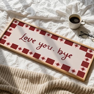 Puede incluir: Un letrero rectangular de madera con las palabras "Love you. bye" en escritura burdeos. El letrero tiene un fondo crema con un borde burdeos de cuadrados y estrellas. Una taza de caf&eacute; y gafas est&aacute;n cerca.