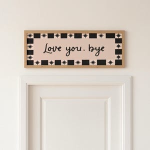 Puede incluir: Un letrero rectangular de madera con las palabras "Love you, bye" en escritura negra sobre un fondo rosa. El letrero est&aacute; enmarcado con un patr&oacute;n de estrellas y cuadrados en blanco y negro.