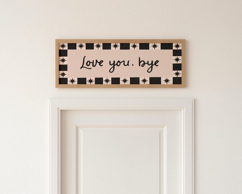 Puede incluir: Un letrero rectangular de madera con las palabras "Love you, bye" en escritura negra sobre un fondo rosa. El letrero est&aacute; enmarcado con un patr&oacute;n de estrellas y cuadrados en blanco y negro.
