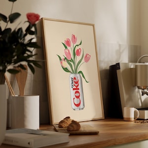 K&ouml;nnte beinhalten: Gerahmter Kunstdruck mit einer Diet Coke-Dose und einem Strau&szlig; rosa Tulpen. Das Kunstwerk hat einen beigen Hintergrund und ist in einem hellbraunen Holzrahmen gefasst. Der Druck wird auf einer Holztheke pr&auml;sentiert.