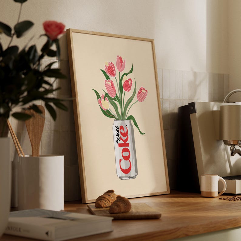 K&ouml;nnte beinhalten: Gerahmter Kunstdruck mit einer Diet Coke-Dose und einem Strau&szlig; rosa Tulpen. Das Kunstwerk hat einen beigen Hintergrund und ist in einem hellbraunen Holzrahmen gefasst. Der Druck wird auf einer Holztheke pr&auml;sentiert.