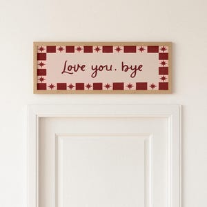 Puede incluir: Un letrero rectangular enmarcado con las palabras "Love you. bye" en un estilo manuscrito. El letrero tiene un fondo rosa claro con un borde rojo y blanco de estrellas y cuadrados. El marco es de color marr&oacute;n claro.