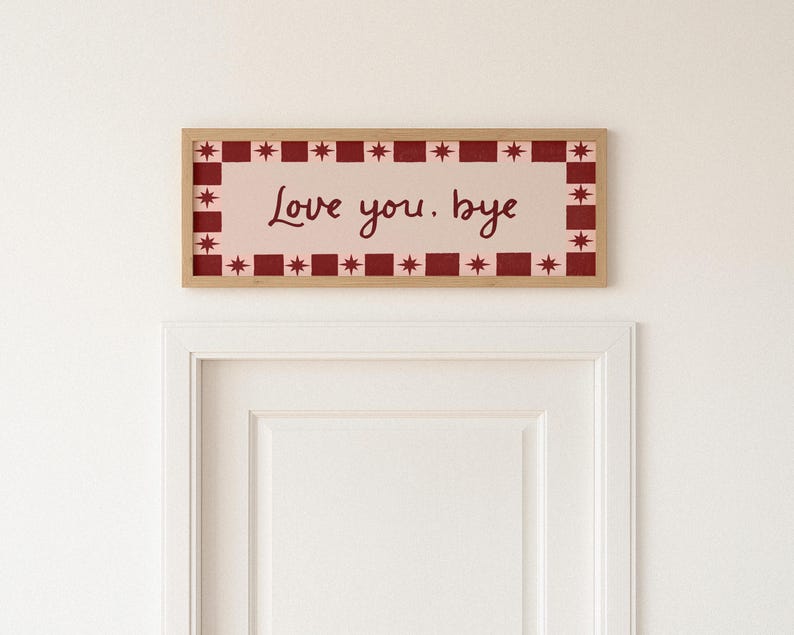 Puede incluir: Un letrero rectangular enmarcado con las palabras "Love you. bye" en un estilo manuscrito. El letrero tiene un fondo rosa claro con un borde rojo y blanco de estrellas y cuadrados. El marco es de color marr&oacute;n claro.