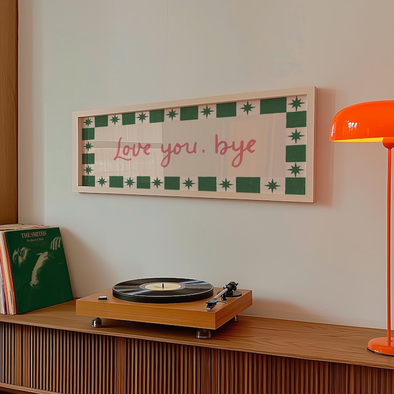 Puede incluir: Una obra de arte enmarcada con las palabras "Love you. bye" en escritura rosa. El arte tiene un borde verde y blanco con estrellas y cuadrados. Un tocadiscos y discos de vinilo est&aacute;n en un mueble de madera debajo.