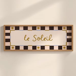 May include: A rectangular framed wall art piece featuring the text le Soleil in gold script. The border consists of a repeating pattern of black and light pink squares, accented with small gold sun icons at the corners and centre points.